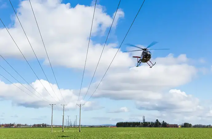 En helikopter som flyger nära marken. I förgrunden en grönskande markyta och i bakgrunden blå himmel med enstaka moln. Intill helikoptern syns elledningar som hänger i luften mellan trästolpar.