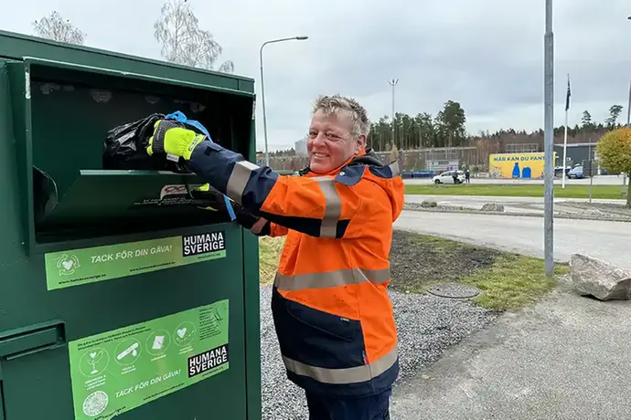 En man i orange jacka lägger in en svart plastpåse i ett grönt skåp. Mannen kollar in i kameran och ler.